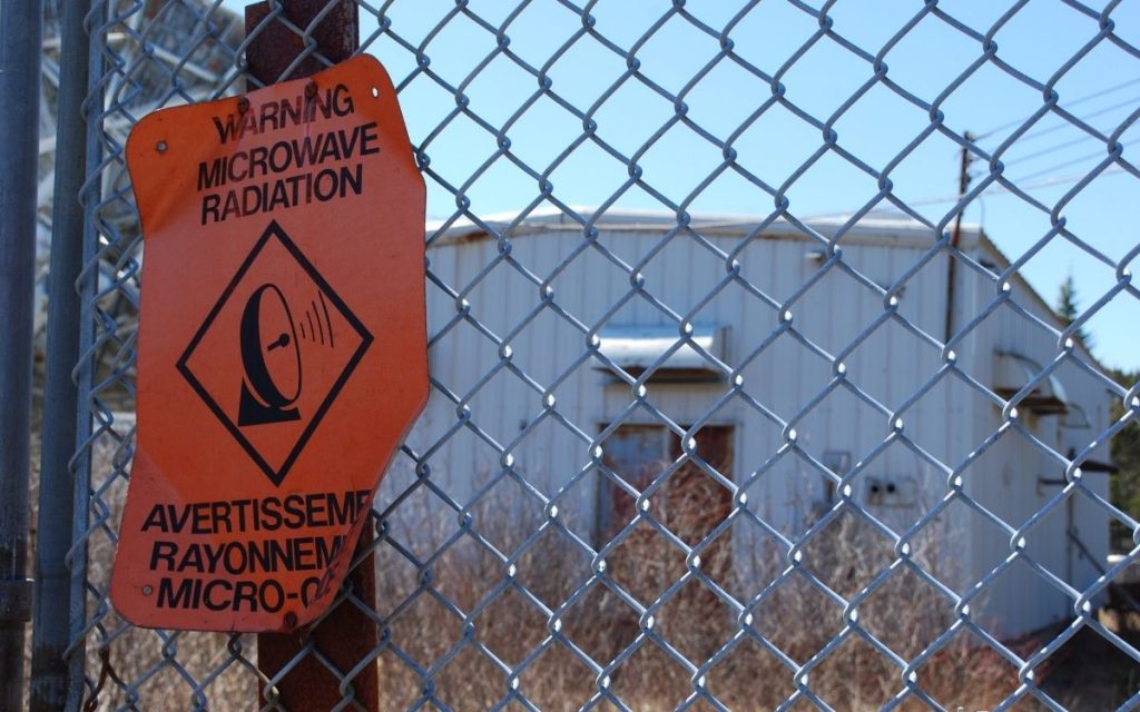 A sign posted on a fence outside a facility reads "Warning microwave radiation".   