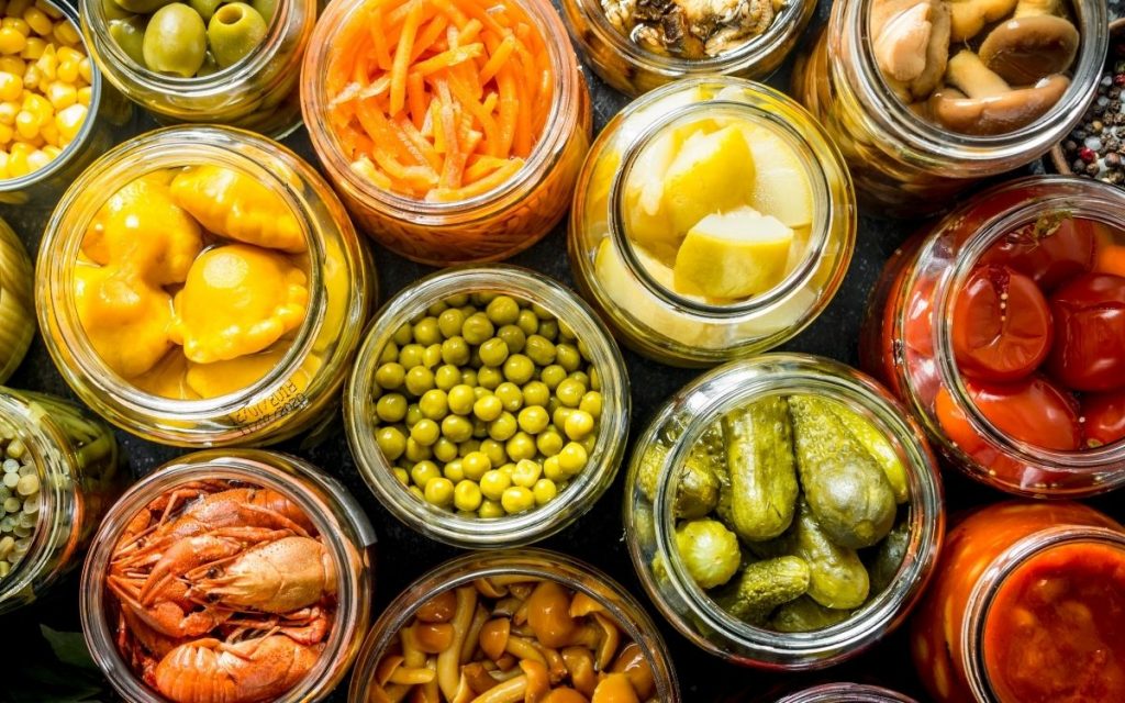Assortment of pickled foods sit with their lids off all in seperate jars.
