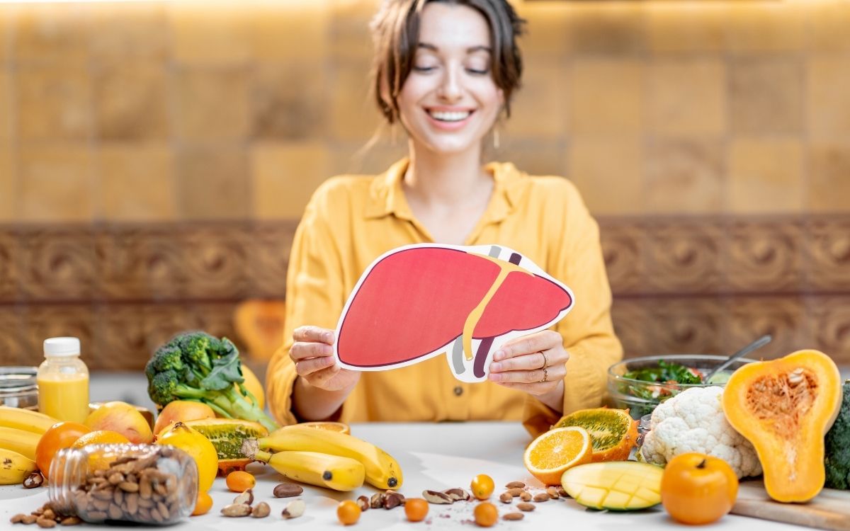 A women holds a picture of a liver in front of multiple foods that are good for the liver.